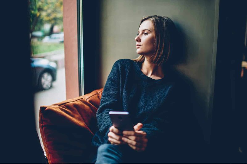 Eine nachdenkliche Frau sitzt am Fenster und hält ein Handy in der Hand
