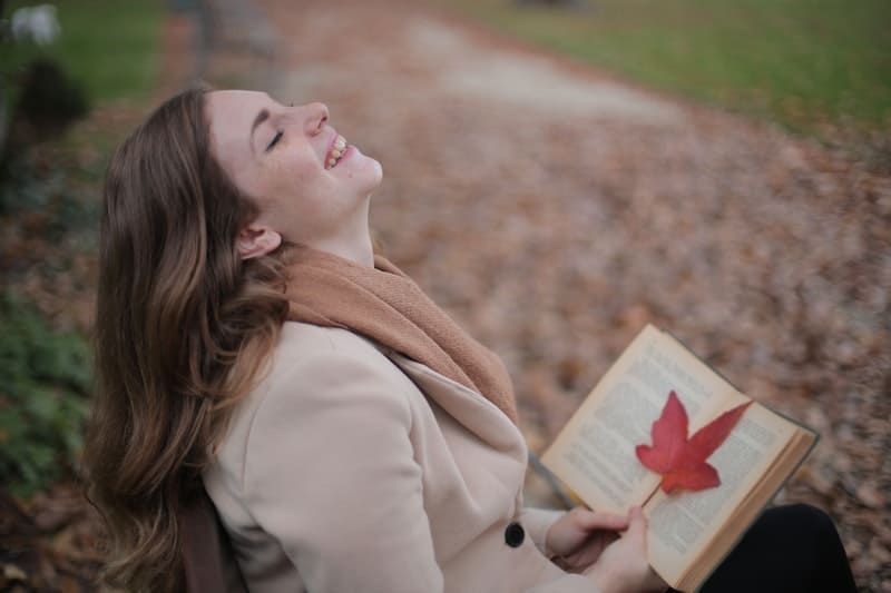 Frau lächelt allein mit einem Buch in der Hand und einem Ahornblatt im Buch