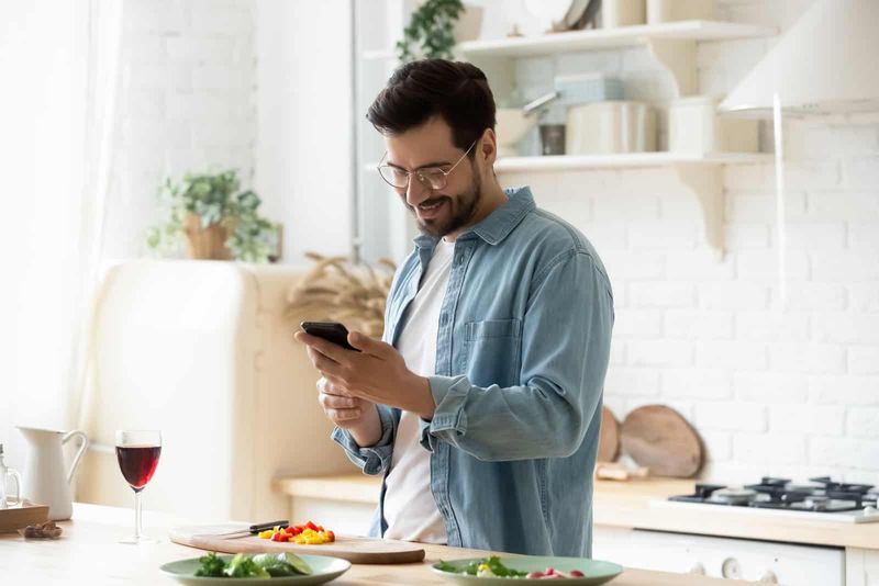 Ein lächelnder Mann mit Brille auf dem Kopf, der in der Küche steht und einen Knopf am Telefon drückt