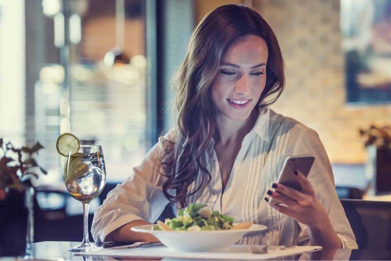 Eine Frau mit langen braunen Haaren sitzt in einem Café und klopft ans Telefon