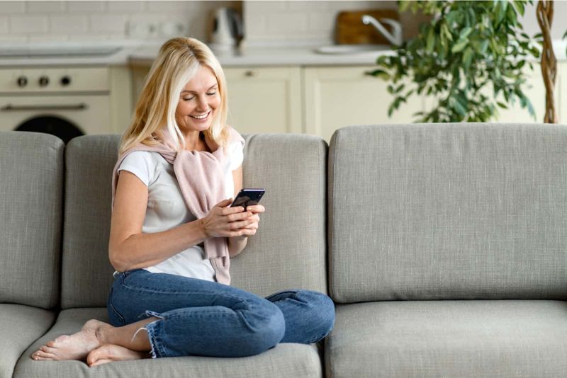 Eine schöne Frau mit blonden Haaren sitzt auf der Couch und schaltet das Telefon ein