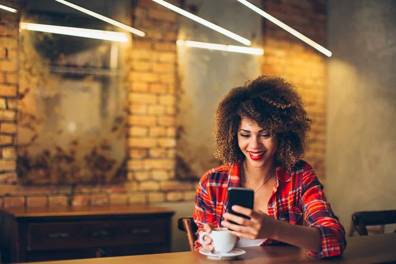 Eine lächelnde Frau mit krausem Haar sitzt in einem Café und tippt am Telefon