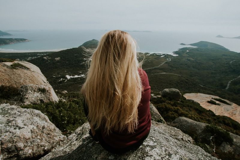 blonde Frau sitzt auf einem Felsen und blickt auf den Ozean