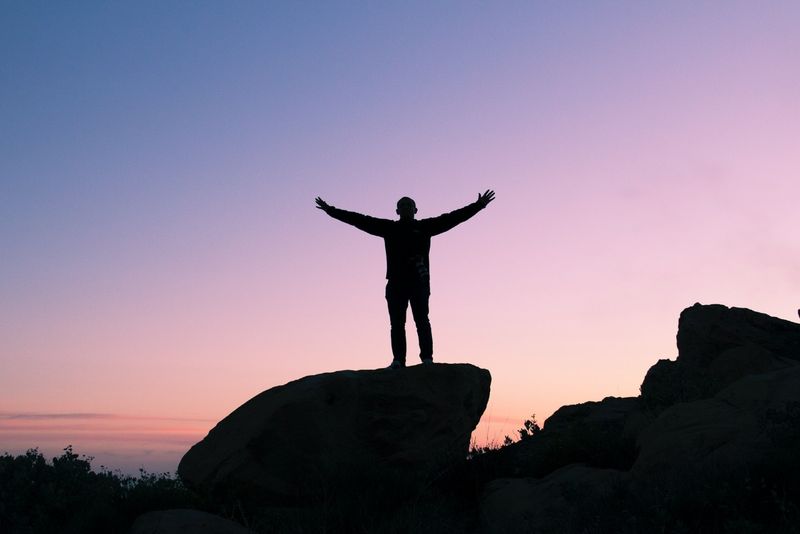 Mann hebt seine Hände, während er bei Sonnenuntergang auf einem Felsen steht