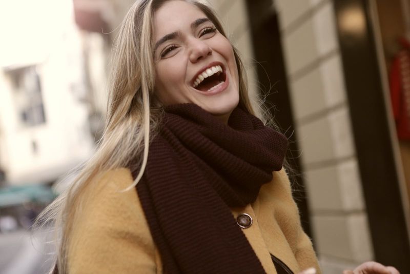 Frau mit braunem Schal lacht, während sie in der Nähe eines Gebäudes steht