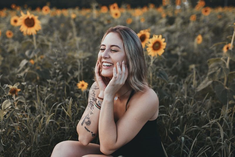 Frau im schwarzen Top lacht, während sie in der Nähe eines Sonnenblumenfeldes sitzt