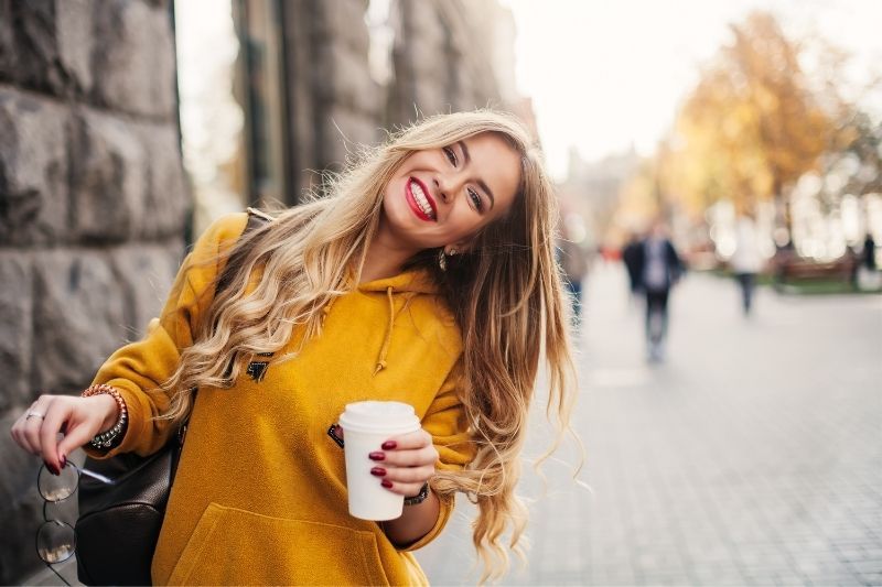 Fröhliche lächelnde Frau, die ihren Kopf leicht nach rechts neigt und mit Kaffee in der Hand im Freien spaziert