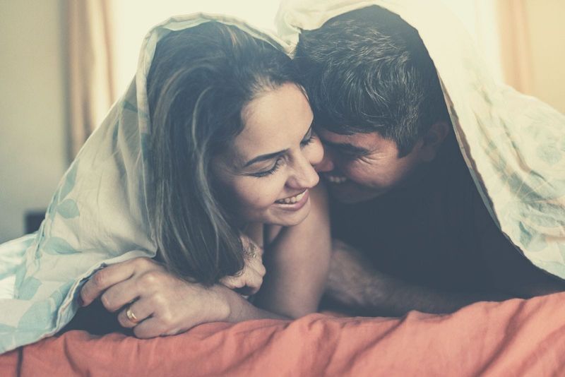 Paar morgens kuschelnd und glücklich, während es mit einer Decke bedeckt im Bett liegt