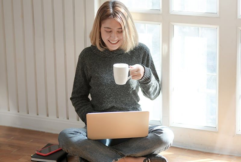 Frau im grauen Pullover trinkt Kaffee, während sie ihren Laptop benutzt