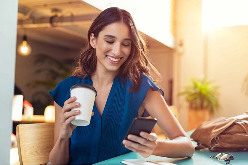 eine junge lächelnde frau sitzt an einem tisch, trinkt kaffee und hält ein telefon in der hand