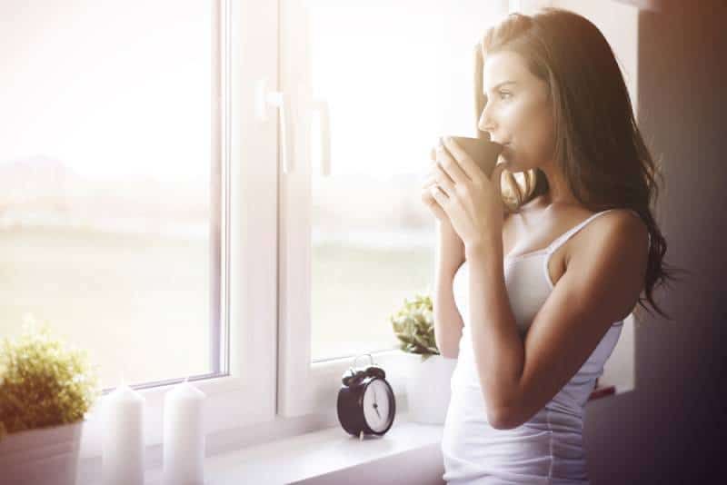 Frau steht morgens vor dem Fenster und trinkt Tee