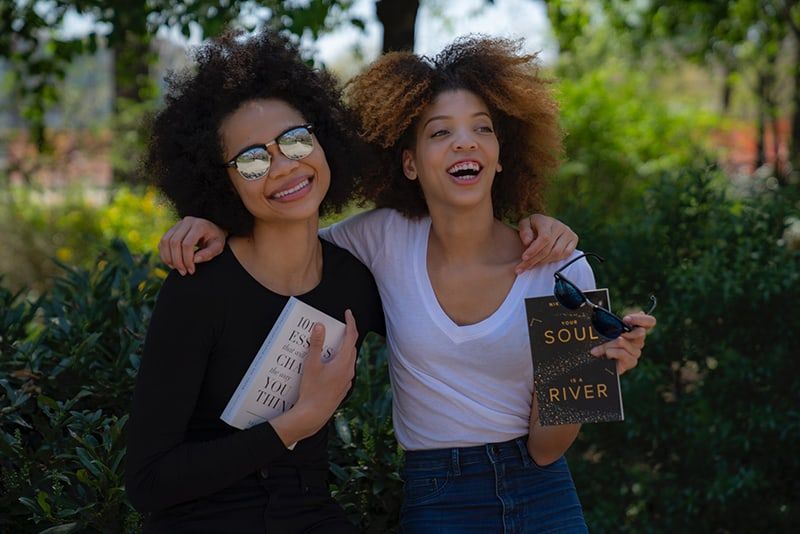 Zwei lächelnde Frauen halten Romanbücher in der Hand, umgeben von Bäumen