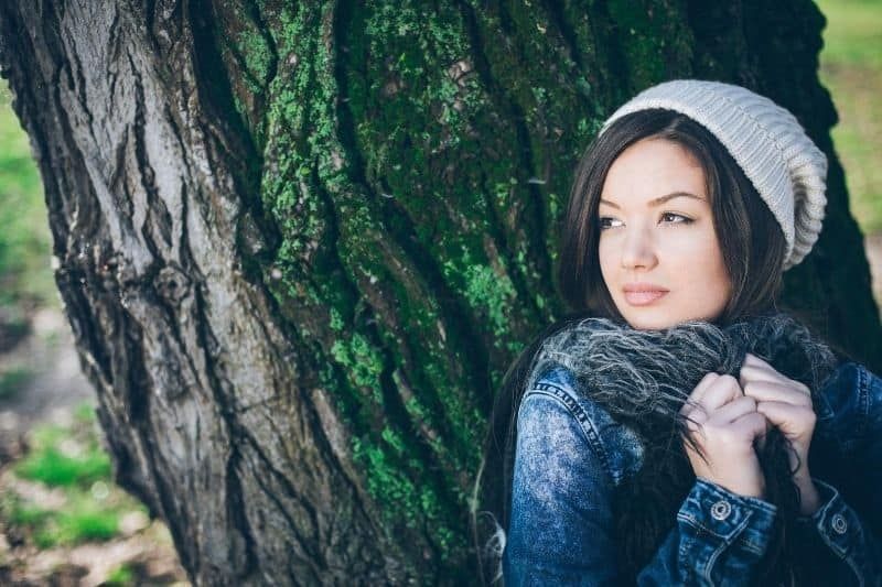 Nachdenkliche Frau versteckt sich hinter dem großen Baum und trägt Jacke und Haube