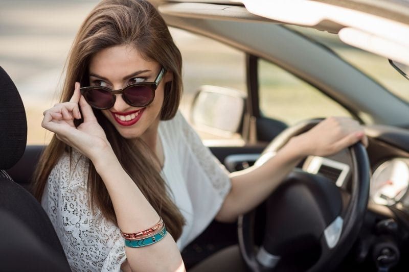 Schöne lächelnde Frau, die in einem von oben nach unten fahrenden Auto fährt und nach hinten schaut und ihre Sonnenbrille hält