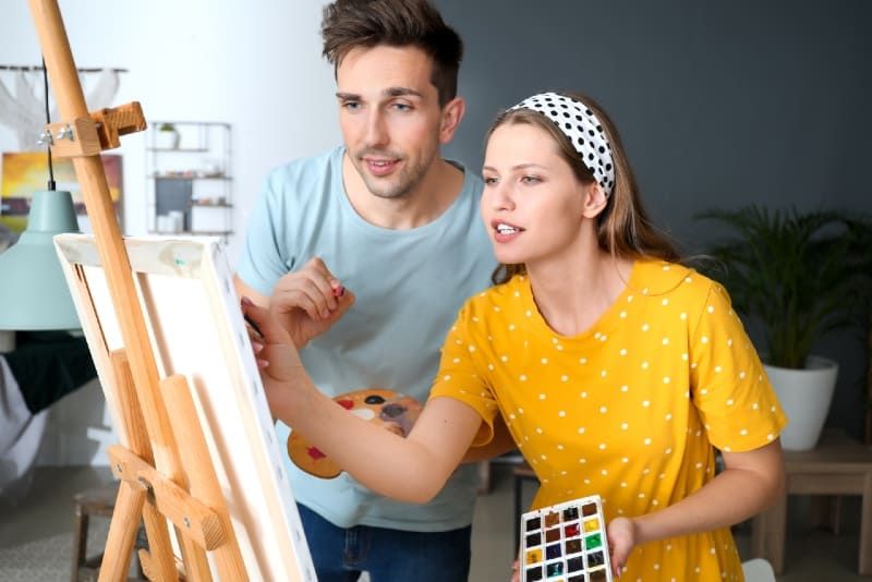 Frau in gelbem Oberteil und Mann malt im Stehen im Innenbereich