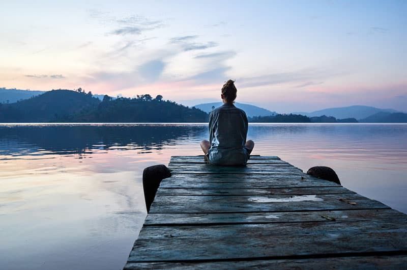 Friedliche Lifestyle-Aufnahme einer Frau, die bei Sonnenuntergang auf dem Dock am Lake Bunyonyi, Uganda, Afrika sitzt.