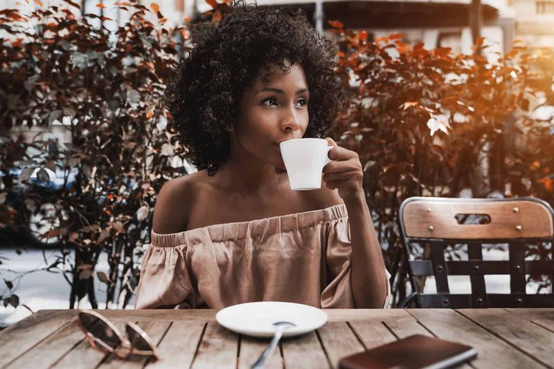 Eine Frau mit krausem Haar sitzt an einem Tisch, trinkt Kaffee und schaut weg