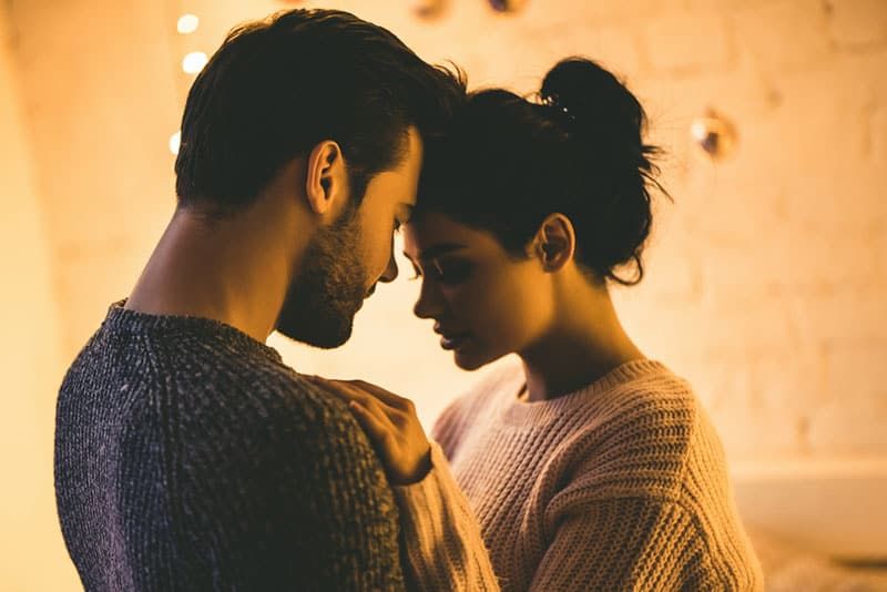 Leidenschaftlich romantisches Paar in Pullovern verbringt Zeit vor Neujahr zu Hause. Zartes Paar umarmt sich vor dem Hintergrund einer glänzenden Girlande.