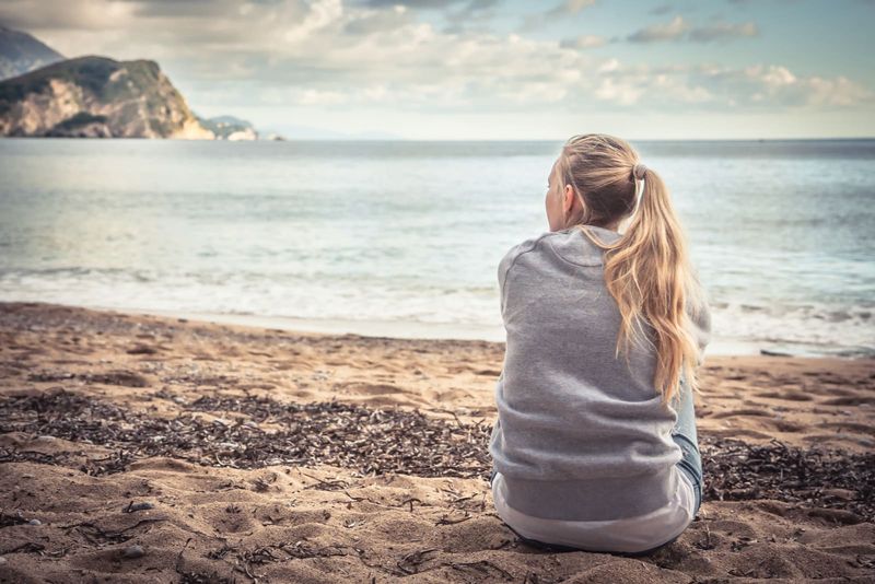 Einsame junge Frau sitzt am Strand und umarmt ihre Knie