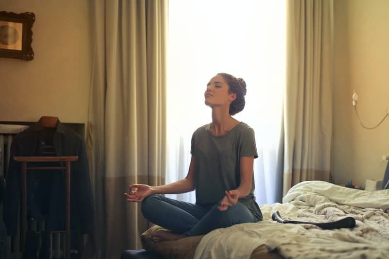 Frau meditiert im Bett im Schlafzimmer mit halboffenen Vorhängen