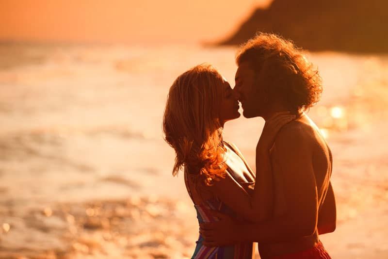 Junge Frau im Bikini küsst ihren Freund am Strand bei Sonnenuntergang. Schönes Paar