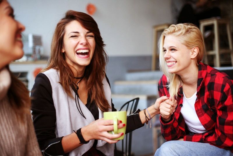 Fröhliche Freundinnen lachen und trinken gleichzeitig Kaffee im Café/Wohnzimmer