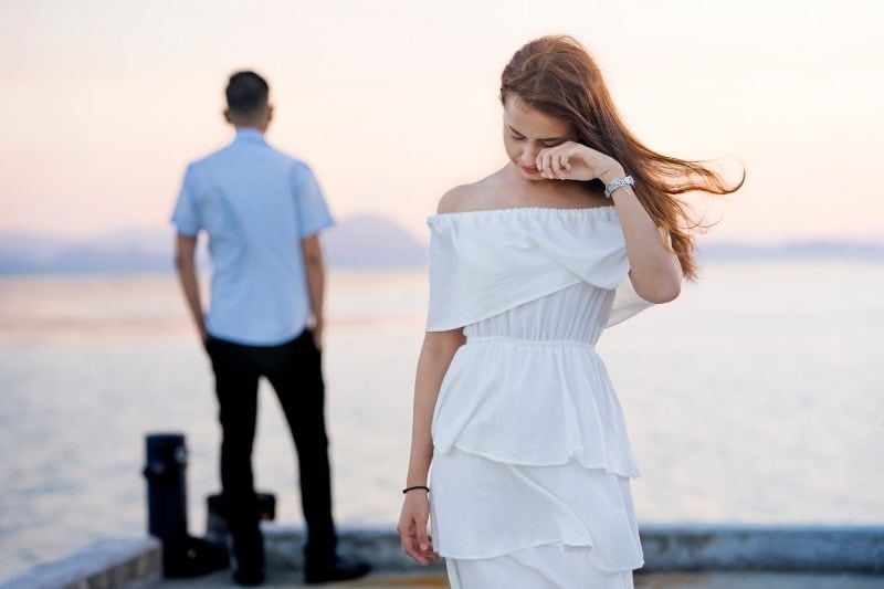junge Frau weint in weißem Kleid und geht von einem Mann weg, der auf ein Gewässer blickt
