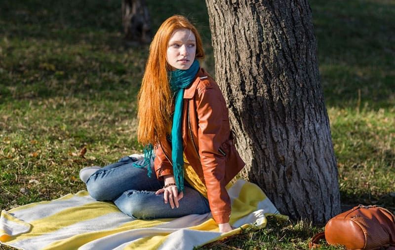 Frau sitzt auf einer Picknickmatte unter einem Baum und blickt in die Ferne und trägt eine braune Jacke