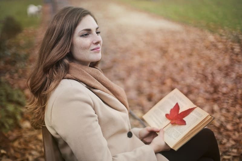 Frau hält ein Buch mit einem Ahornblatt und sitzt im Herbst auf der Bank im Park