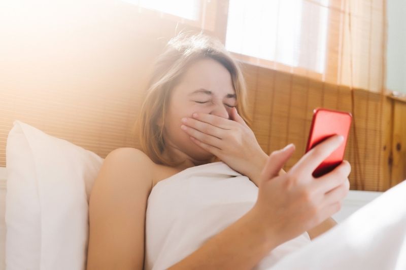 Frau liegt im Bett und niest, während sie ihr Smartphone hält