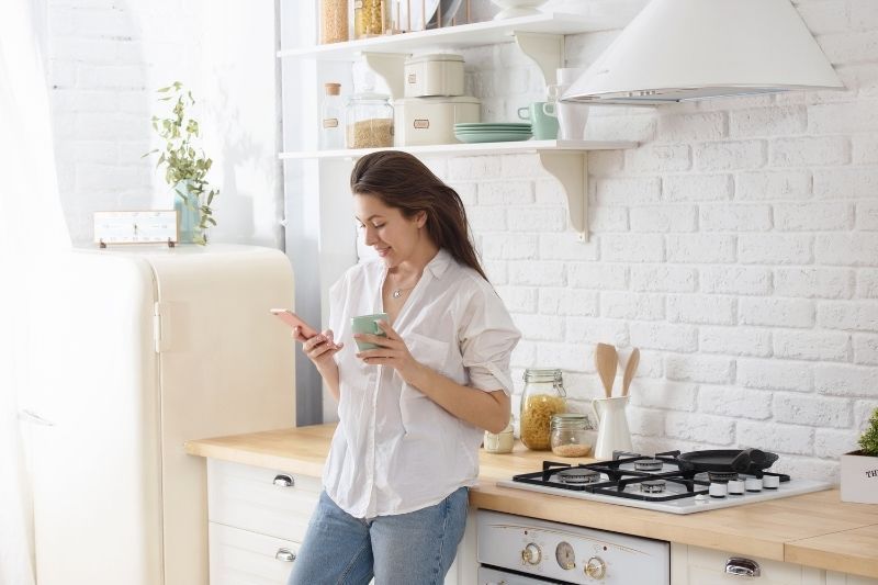 Lächelnde Frau steht in der Küche, hält ein Mu in der Hand und liest eine SMS auf ihrem Mobiltelefon