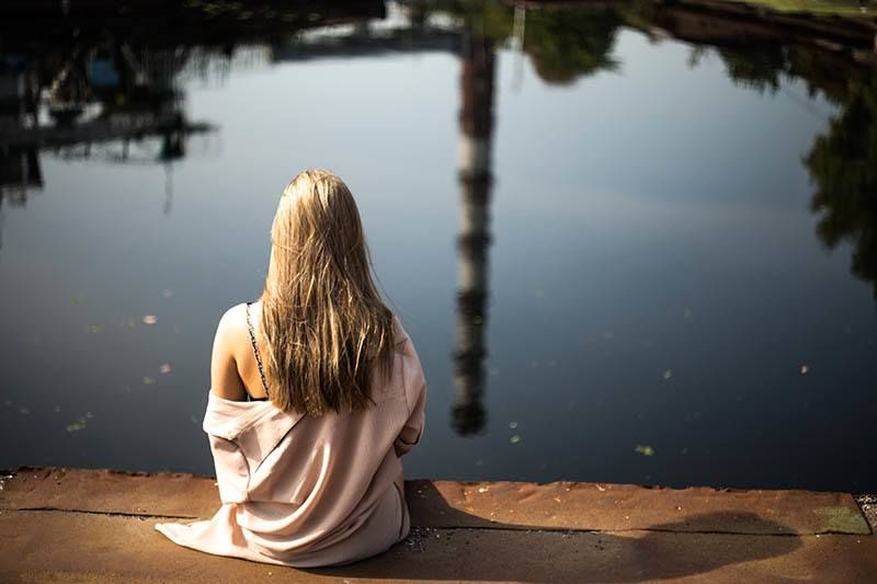 Rückansicht einer blonden Frau, die auf einem Holzsteg vor dem See sitzt