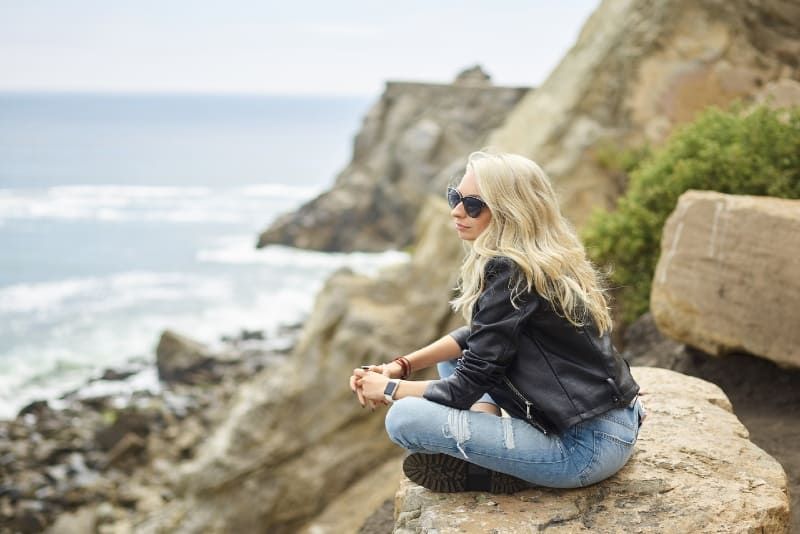blonde Frau mit Sonnenbrille sitzt auf einer Klippe