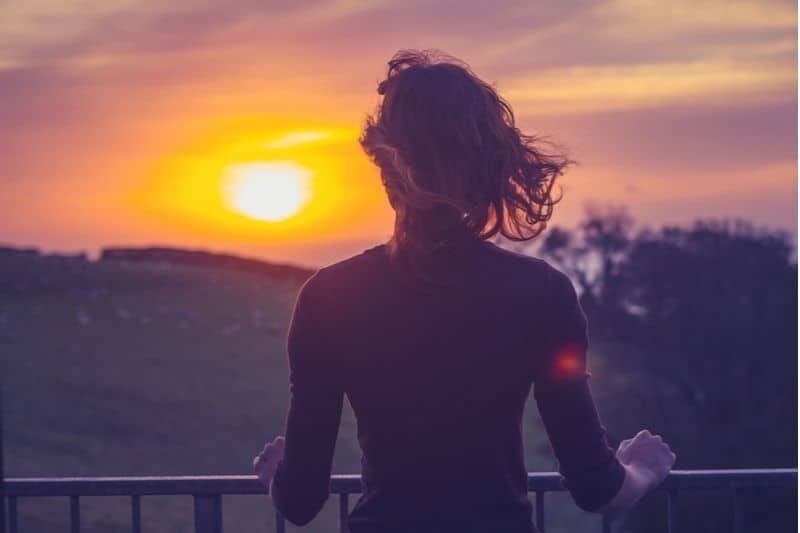 Frau beobachtet Sonnenaufgang/Sonnenuntergang und hält sich am Geländer fest