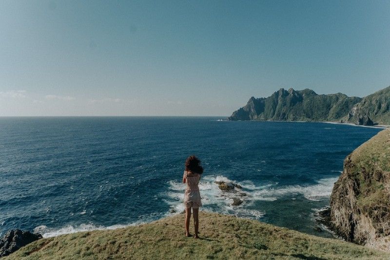 Frau im weißen Oberteil, die auf einer Klippe steht und auf das Meer blickt