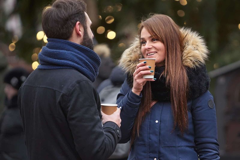 Ein lächelnder Mann und eine lächelnde Frau stehen draußen, halten Tassen Kaffee in der Hand und reden