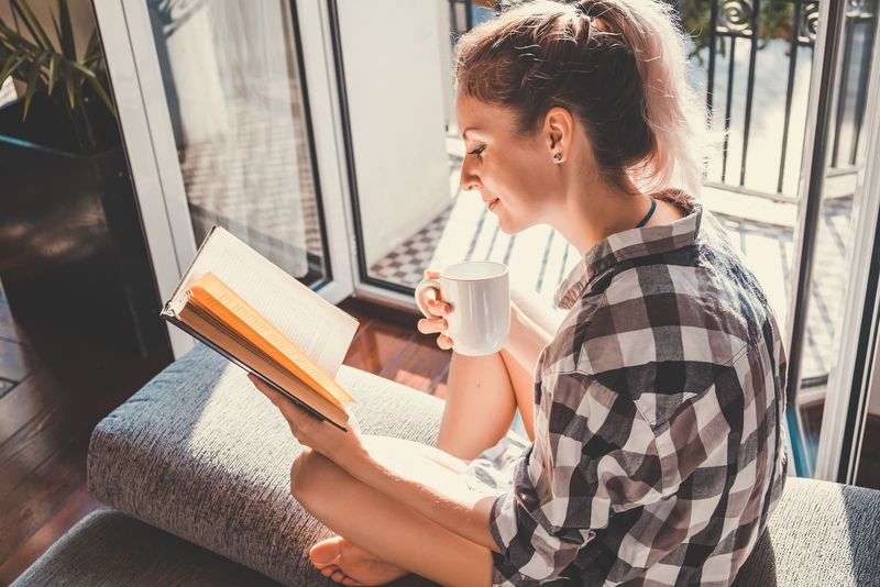hübsche Frau, die am geöffneten Fenster sitzt, Kaffee trinkt und ein Buch liest