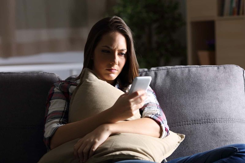 Besorgte Frau schaut auf ihr Mobiltelefon, während sie auf der Couch sitzt