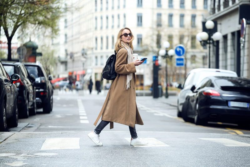 Eine Frau in einem braunen Mantel mit einem Telefon in der Hand geht die Straße entlang