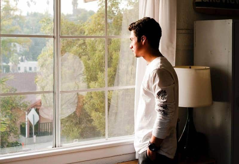Mann im weißen T-Shirt steht neben dem Fenster und schaut hinaus