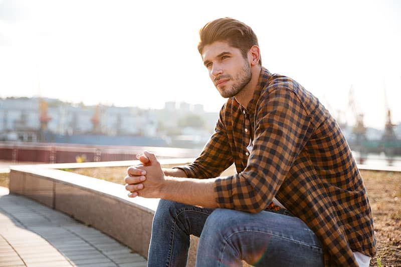 Nachdenklicher junger Mann im karierten Hemd, der draußen sitzt und nachdenkt