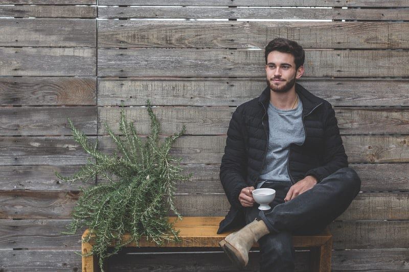 Mann sitzt auf einer Holzbank in der Nähe einer Bretterwand und neben sich steht eine Pflanze, die eine Tasse hält