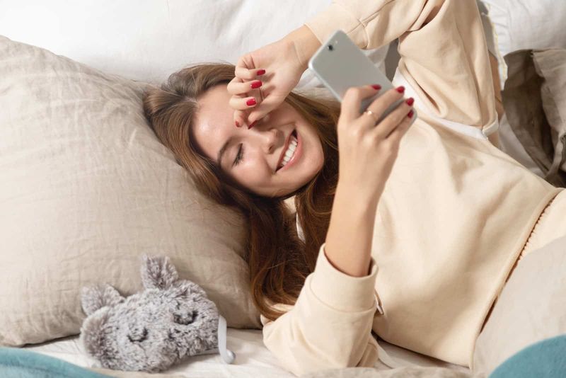 Eine lächelnde Frau liegt im Bett und schaut auf das Telefon