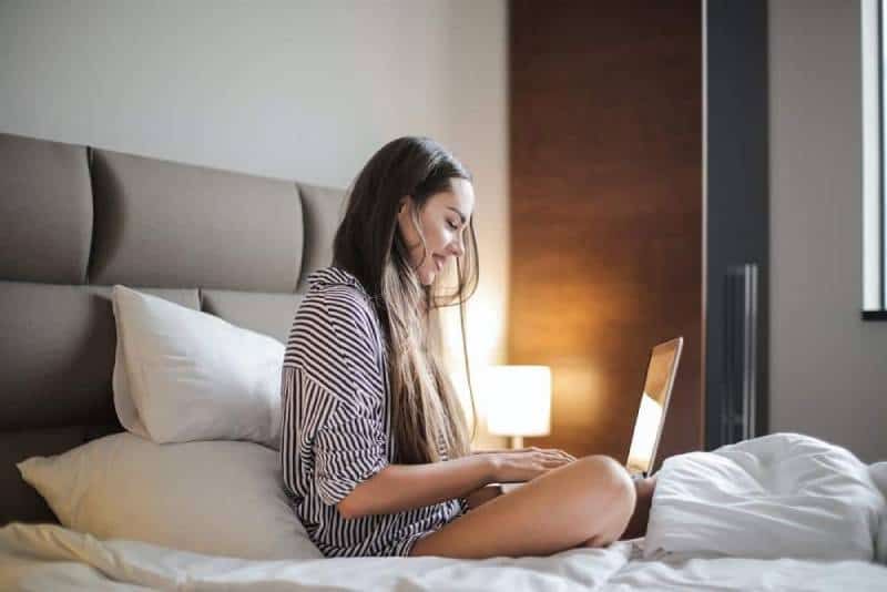 lächelnde Frau in einem schwarz-gestreiften Oberteil, die auf einem Bett sitzt, während sie einen Laptop benutzt