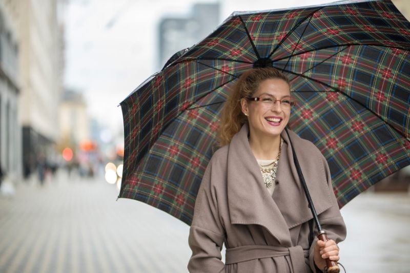 Lächelnde Frau mit Regenschirm, die in einem langen Trenchcoat die Straße entlang geht