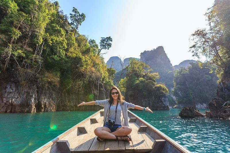Eine Frau sitzt auf einem Boot und segelt durch ein Gewässer inmitten großer Felsklippen und breitet vor Glück die Arme aus