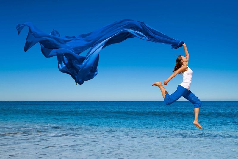 Frau springt vor Freude in der Nähe des blauen Meeres und trägt ein blaues Tuch, das vom Wind geblasen wird