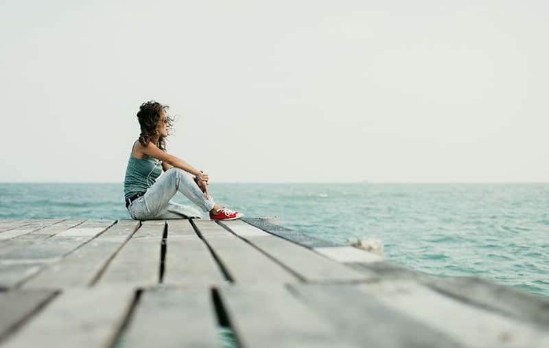 Frau am Meer sitzt auf einer Holzplattform und trägt Turnschuhe