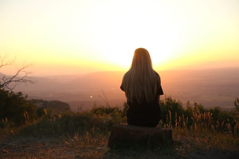 Frau, die den Sonnenuntergang betrachtet, der auf einem Felsen auf einem Berg sitzt