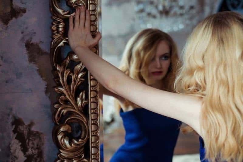 Eine Frau, die in einem blauen Kleid in den Spiegel blickt und die Hände am Rand des Spiegels hält, denkt nach